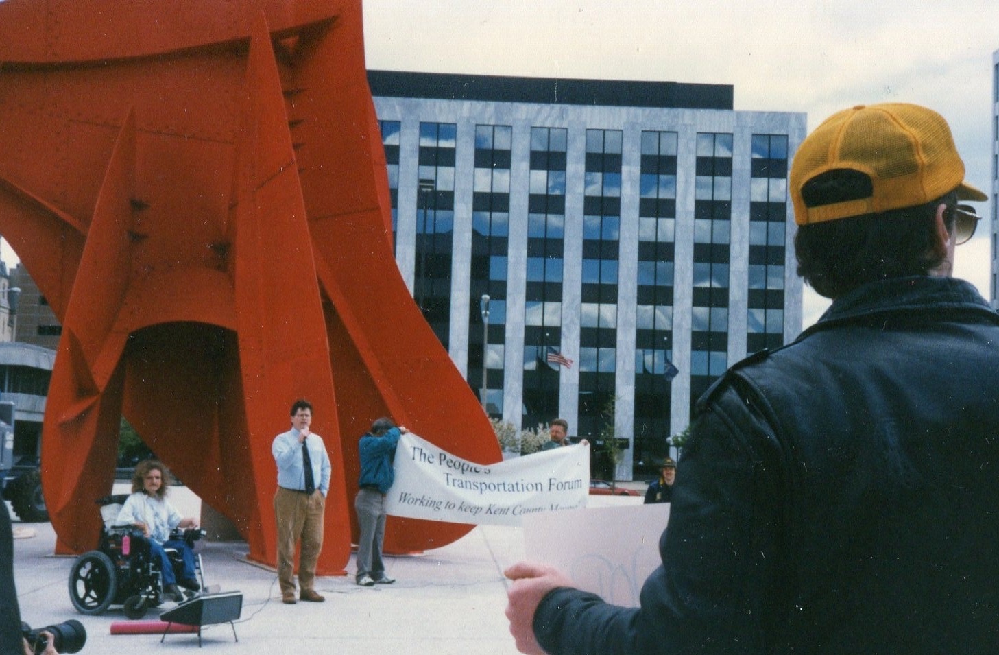 A younger Dave Bulkowski speaking at The People's Transportation Forum near the Calder sculpture in downtown Grand Rapids, with a rider in a wheelchair beside the stage.